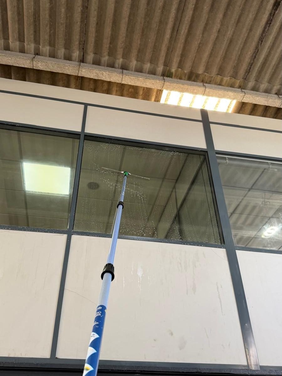 Vara telescópica com cabeça de lavagem encostada a um pano de vidro alto, ainda molhado, no interior de um armazém