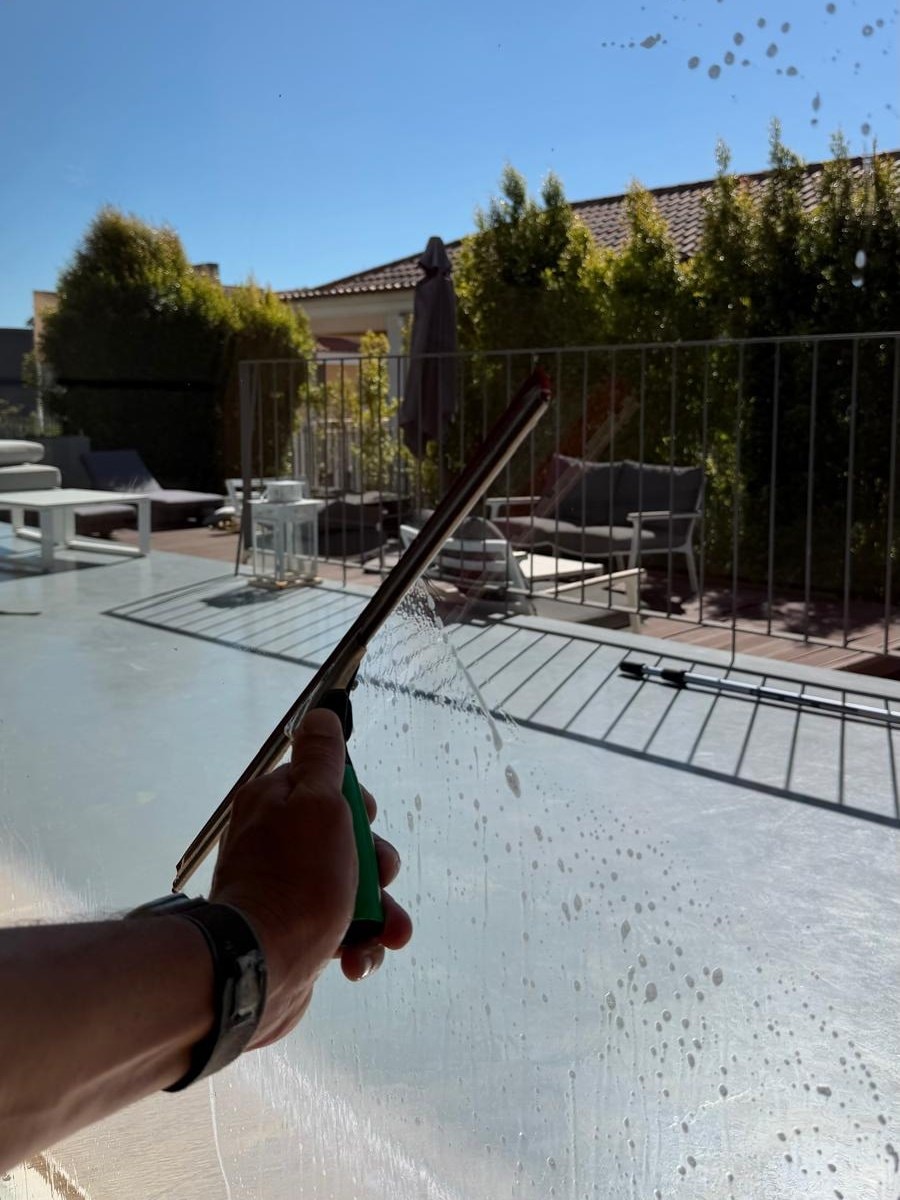 Mão a segurar o rodo contra um pano de vidro ainda molhado, com o terraço e a sebe atrás